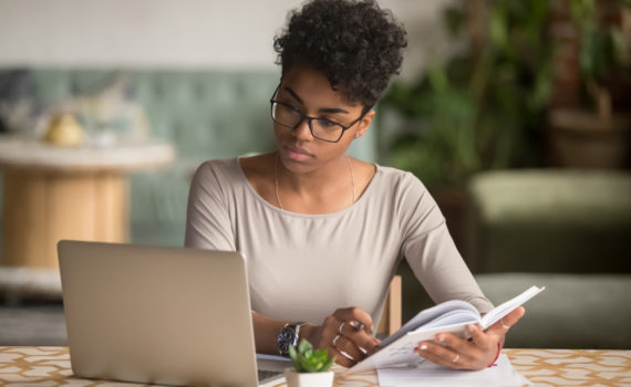 Focused young african american businesswoman or student looking at laptop holding book learning, serious black woman working or studying with computer doing research or preparing for exam online