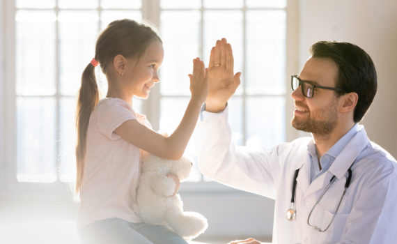 Side view happy little preschool girl giving high five to male doctor at meeting in hospital. Smiling small patient celebrating successful treatment finish with general practitioner at checkup.